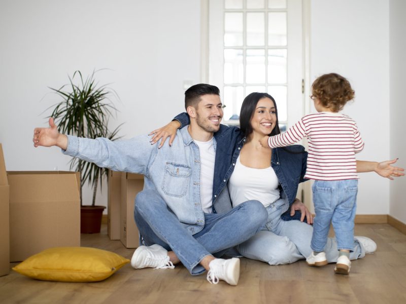 Happy young family with little son having fun at new home after moving, smiling millennial parents playing with their child while sitting on floor among cardboard boxes, opening arms for embrace